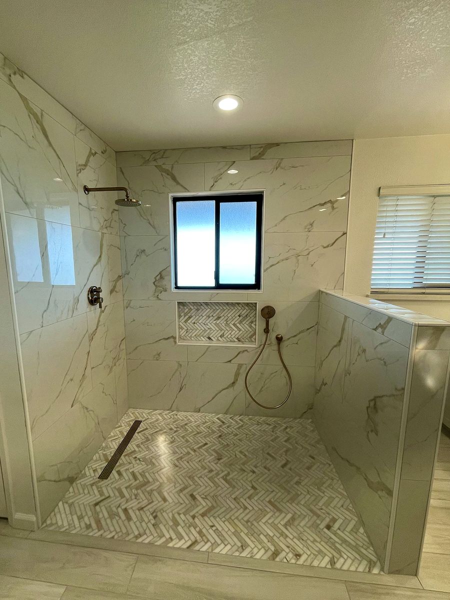 Curbless walk-in shower with marble-look wall tile, a herringbone tile floor, a linear drain and a brass rain head and handheld