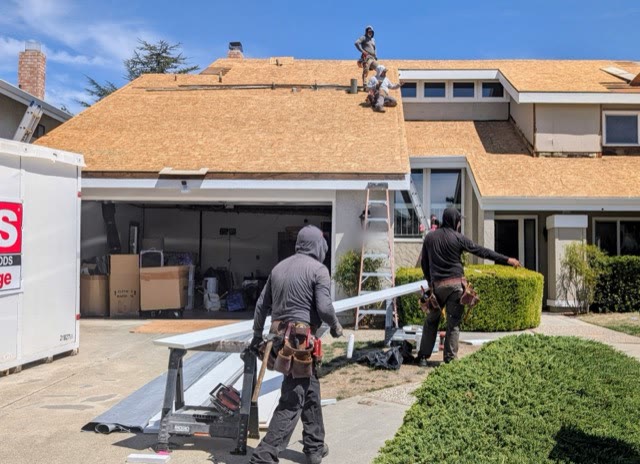 Two-story house with the old wood shake stripped off and new plywood sheathing installed across the roof, crew working on the roof and in the driveway